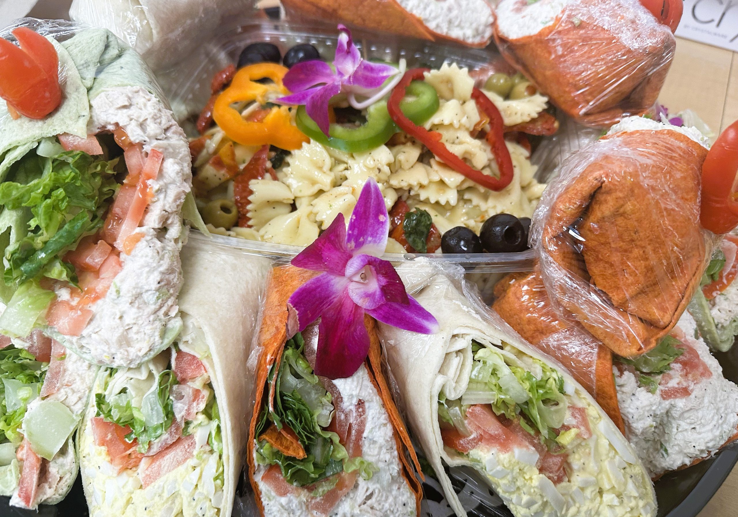 A platter featuring various wrapped sandwiches with cream cheese, lettuce, and tomato, surrounding a pasta salad with olives and sliced bell peppers, garnished with an orchid.